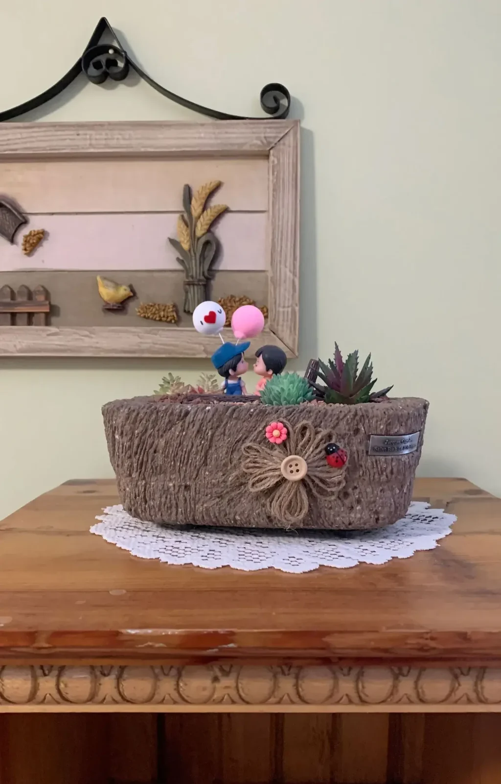 Decorative hypertufa pot with succulents and romantic figurines holding balloons, on a wooden table.
